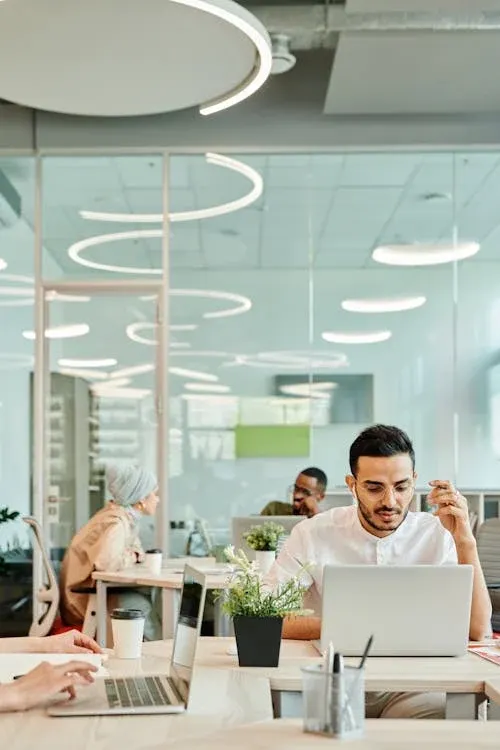 A man working at a laptop in a bright open-plan office, colleagues at desks behind him