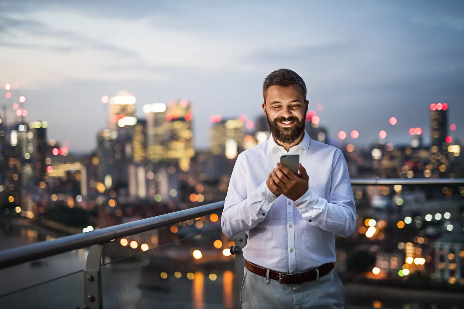 A man in a white shirt looking at his phone on a balcony, with a lit city skyline behind him