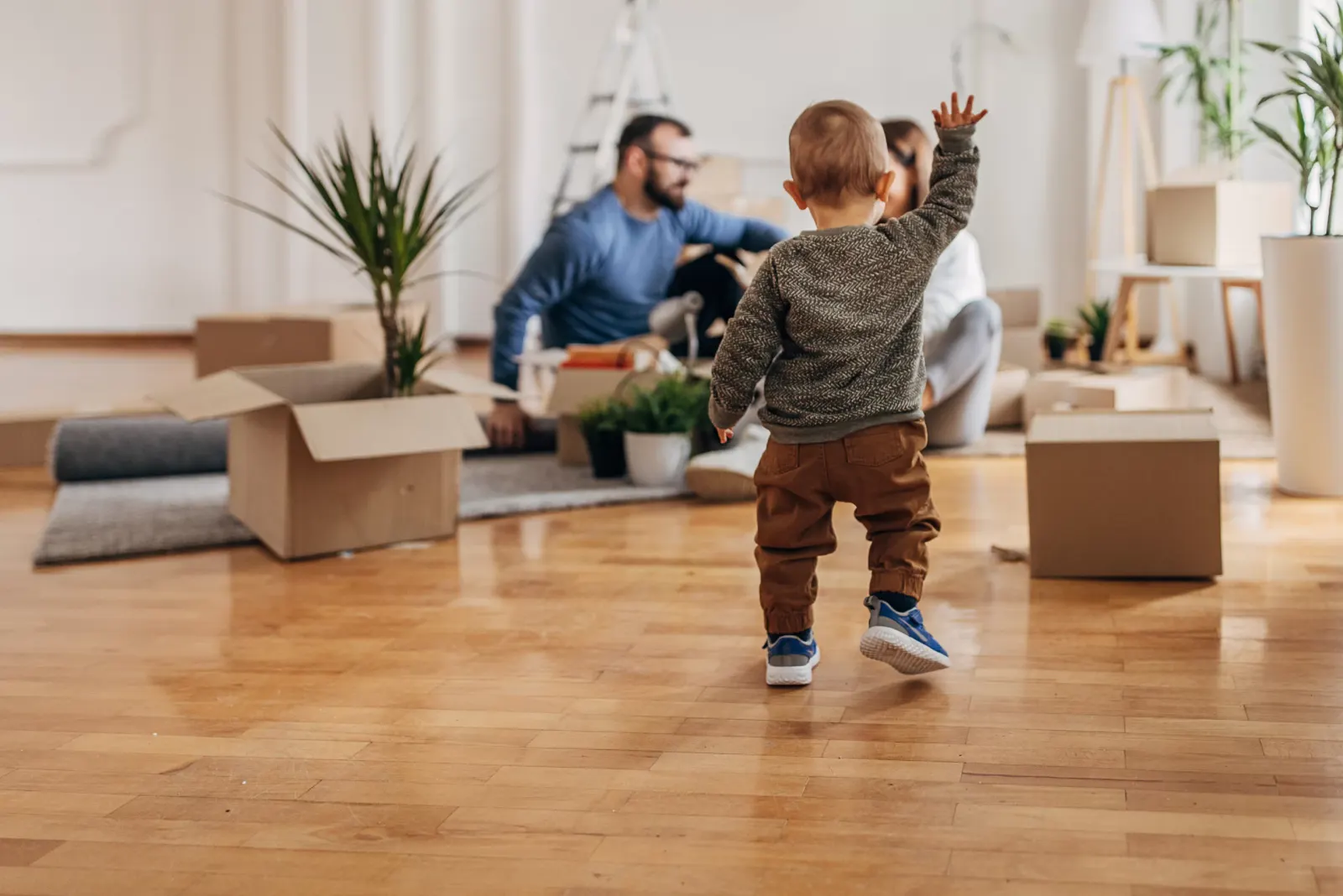 A toddler walking across a wooden floor among packing boxes as two adults unpack behind