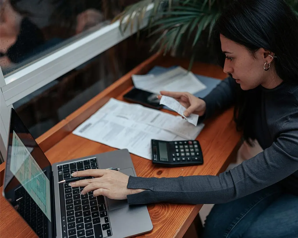 A woman at a desk working through printed statements with a calculator and laptop