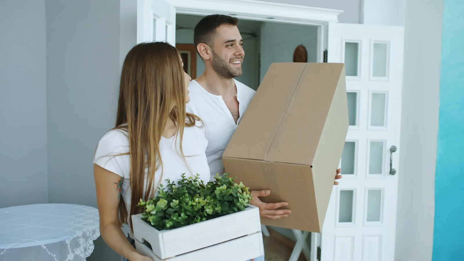 A couple carrying a cardboard box and a plant through a front door